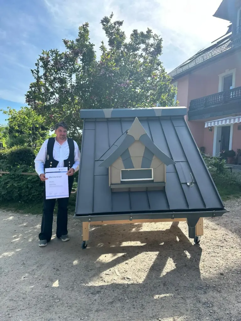 Spenglermeister Marcel Lautz in Zunftkleidung mit seinem Meisterbrief neben einem Dachmodell mit Stehfalzdeckung und verzierter Gaube
